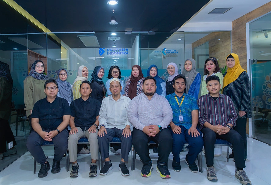 Group of colleagues posing for a team photo in a modern office lobby with glass walls and branding in the background; mix of men and women, several women wearing hijabs, all in business casual attire.