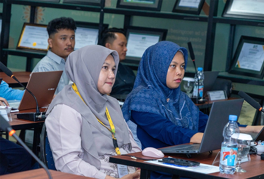 Two women wearing hijabs sit at a conference table with laptops, attentively listening in a meeting; certificates visible on the wall in the background.
