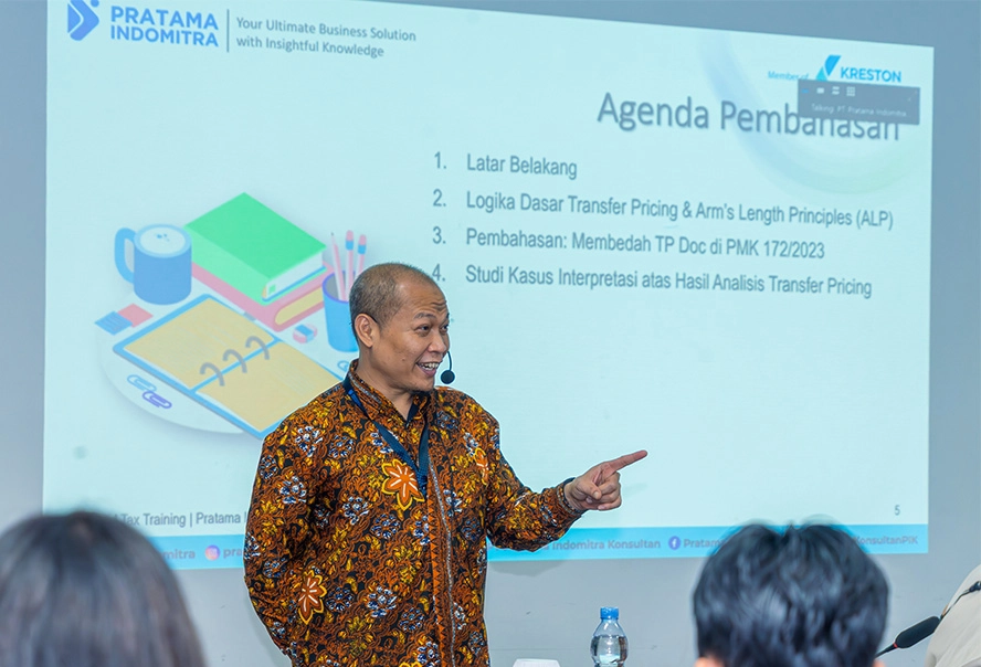 Speaker wearing batik shirt presenting in front of a slide titled 'Agenda Pembahasan' with an agenda list and company logos in the corner.